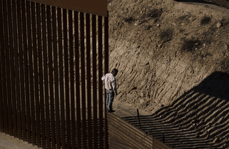 A man stands at a tall fence, looking at the long shadow it casts on the oppposite side.